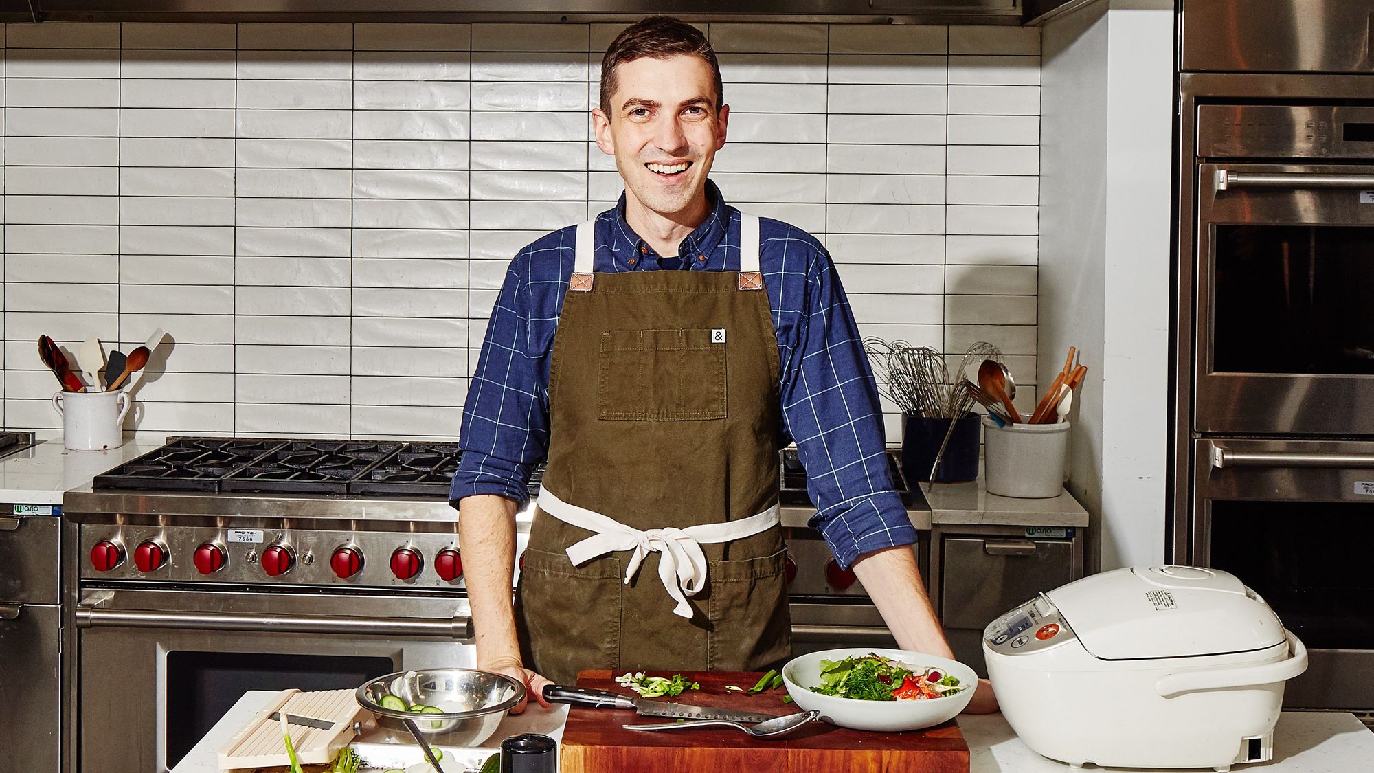 Chris Morocco from Bon Appetit wearing a blue checked shirt and a khaki green apron in front of a counter with a bowl of salad in the BA test kitchen