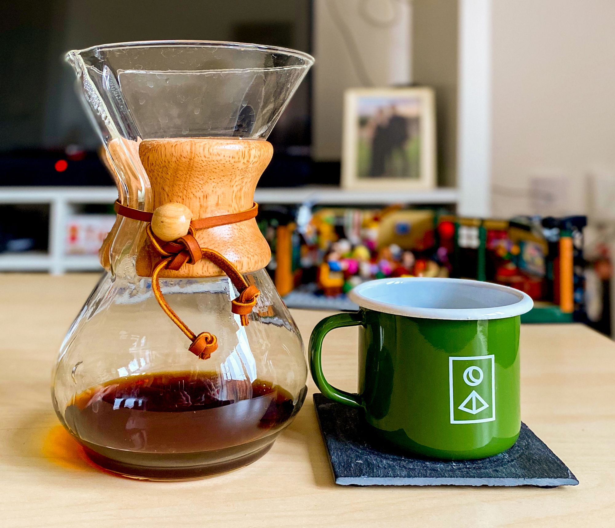 A Chemex coffee dripper with a small amount of coffee left in the bottom and a green enamel mug on a slate coaster, both placed on a wooden coffee table