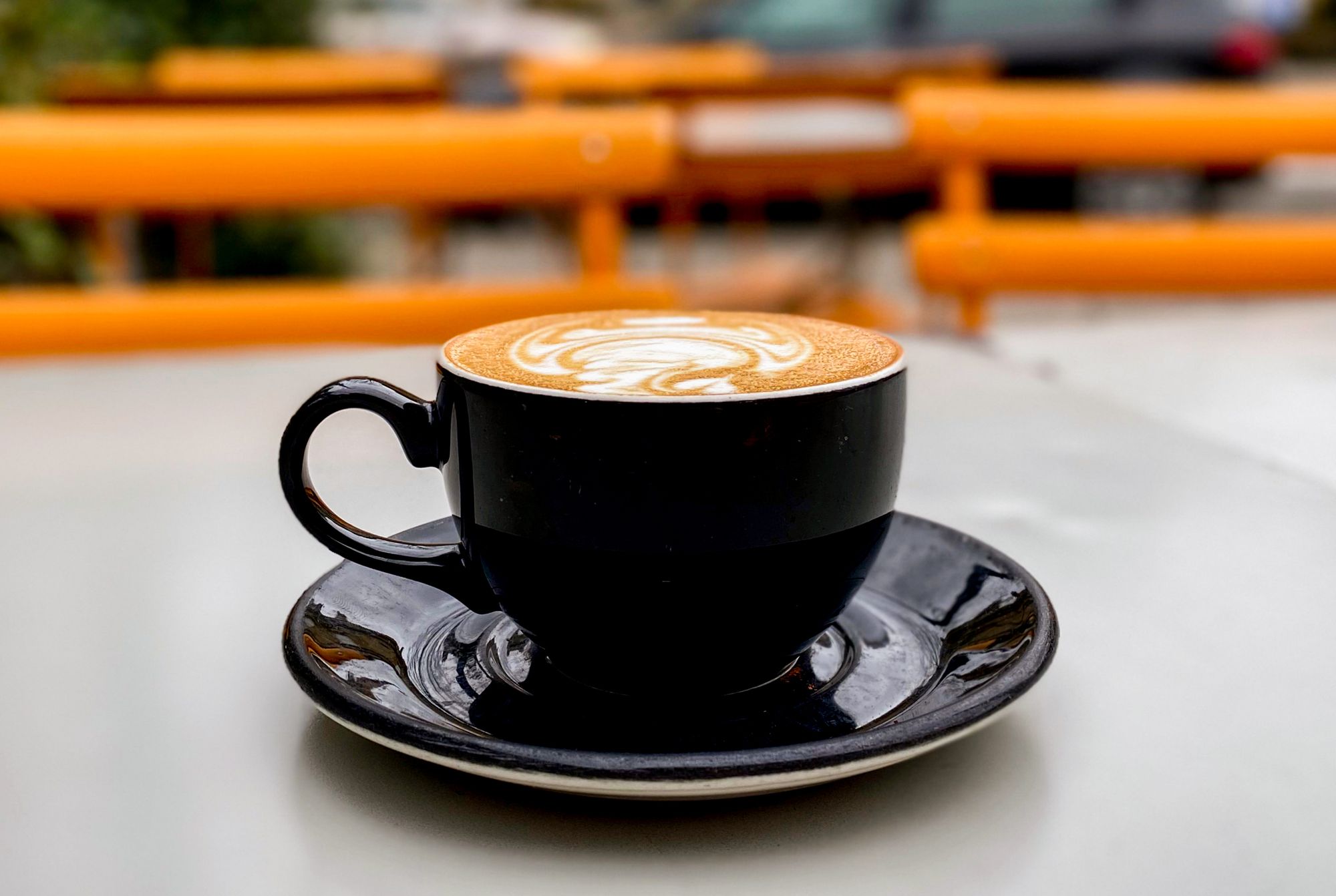 A black ceramic coffee cup with a flat white in it and latte art on a table outside