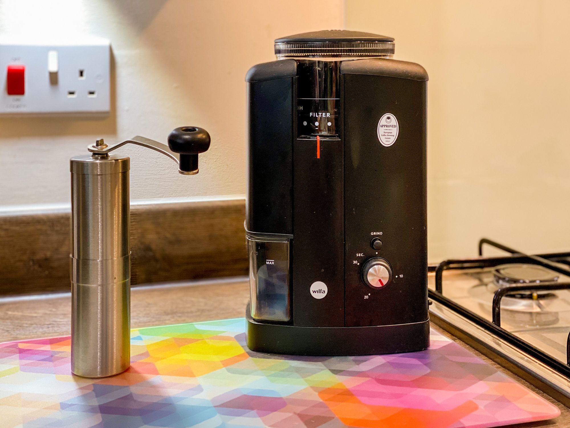 The Wilfa Svart electric burr grinder and a Portex tall hand burr grinder on a colourful glass tray placed on a kitchen countertop