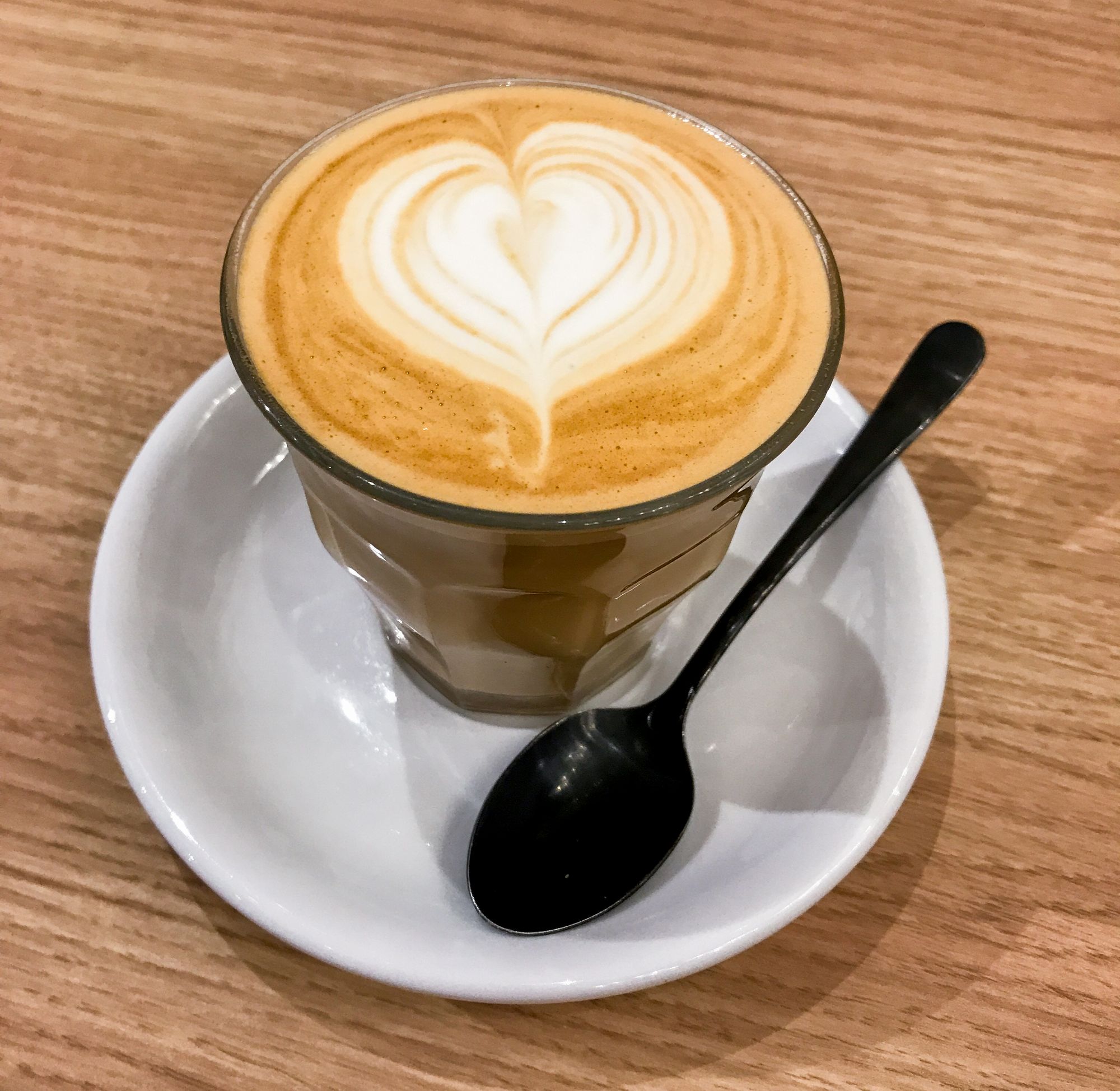 A latte with heart-shaped latte art in a small glass sitting on a white ceramic saucer with a black metal spoon placed on a wooden table