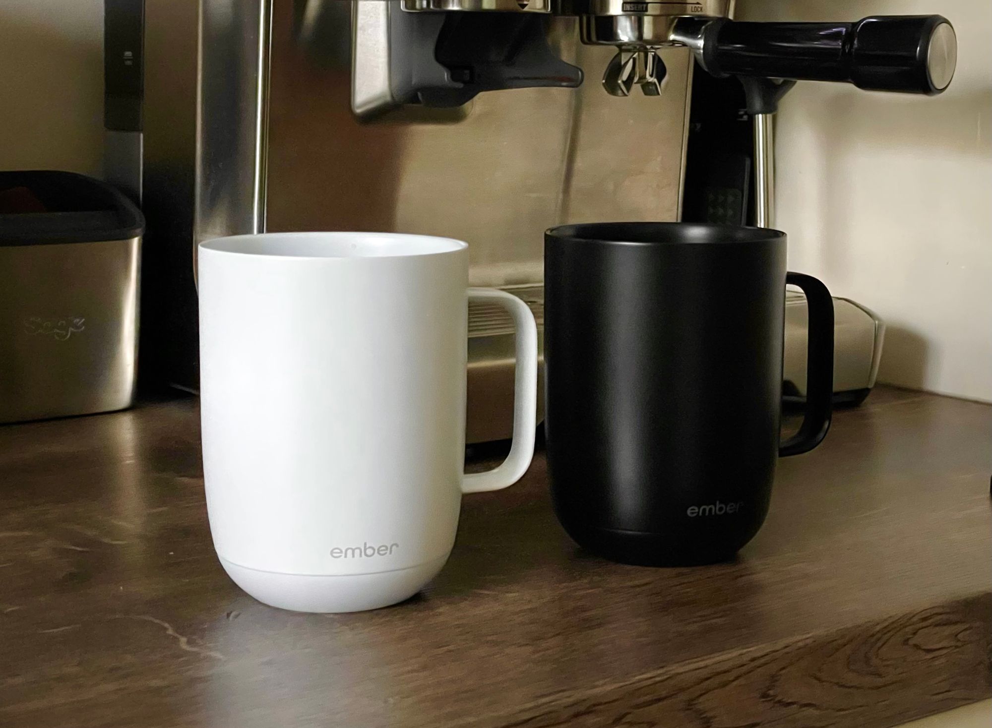Two Ember mugs, one black and one white, on a brown wooden kitchen counter in front of an espresso machine