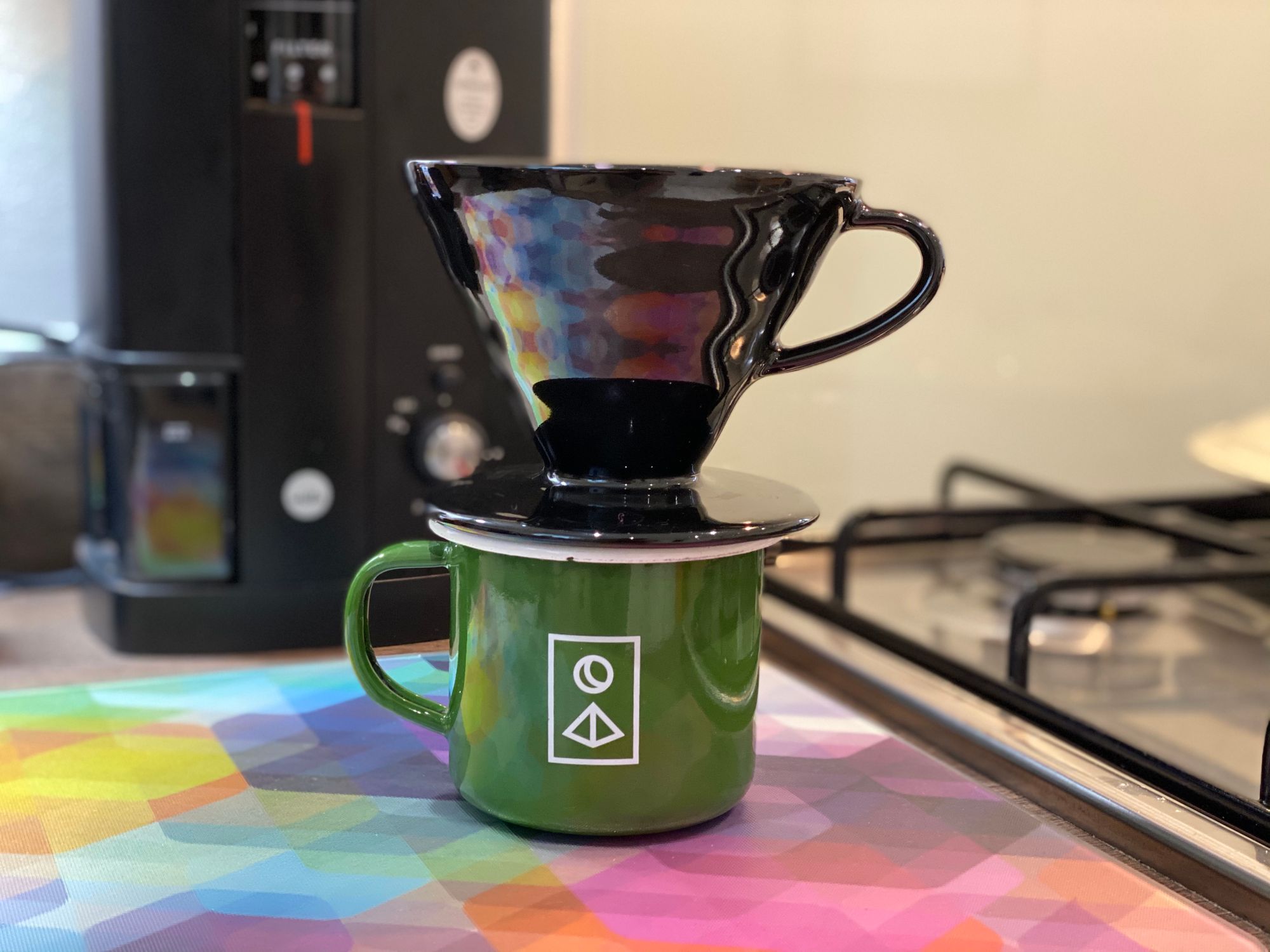 A black ceramic V60 dripper on top of a green enamel Forestry mug on a brightly coloured worktop saver in a kitchen