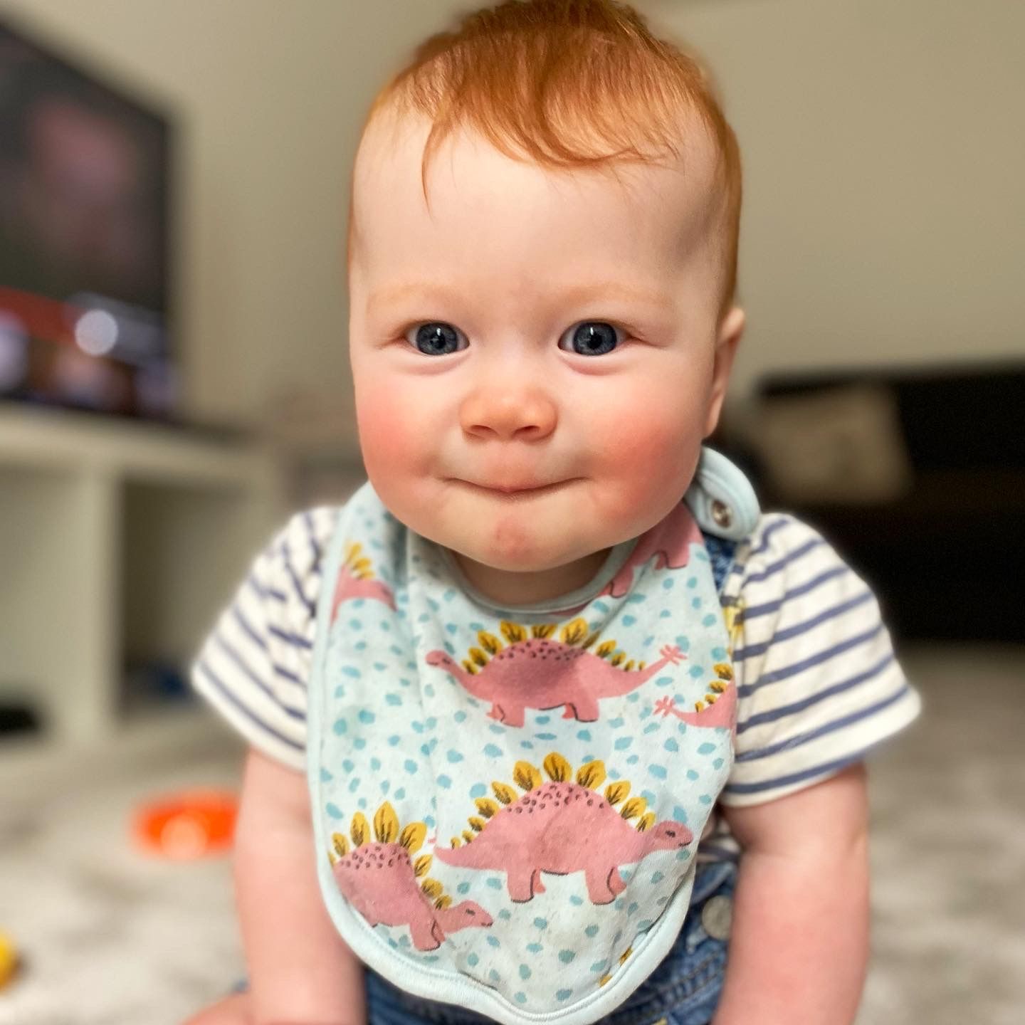 An 8 month old baby with red hair and grey-blue eyes looking at the camera with a coy face