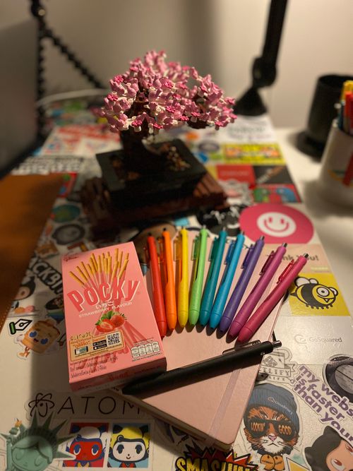 A pink Leuchtterm notebook on a sticker covered desk, topped with rainbow coloured pens and a box of strawberry Pocky, in fr