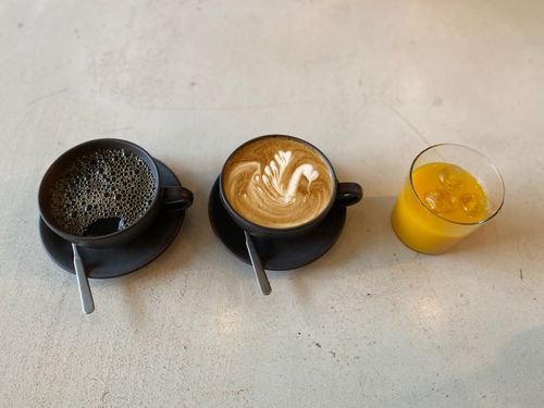 Two cups and saucers of coffee, one black and one latte, next to a glass of orange juice on a concrete table top